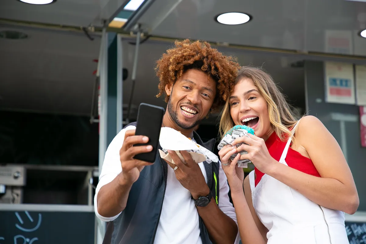 Dos amigos ríen y se toman una selfie con el celular mientras comen en un food truck